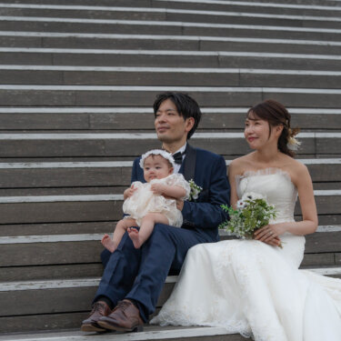 広い木の階段に腰かけて赤ちゃんを膝に抱く新郎と、ブーケを持って寄り添う花嫁。家族写真としての洋装前撮り