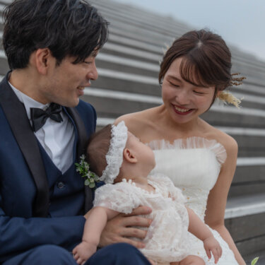 レースのベビードレスの赤ちゃんを抱く花嫁を、笑顔でのぞき込む蝶ネクタイの新郎。親子三人で撮るフォトウェディング