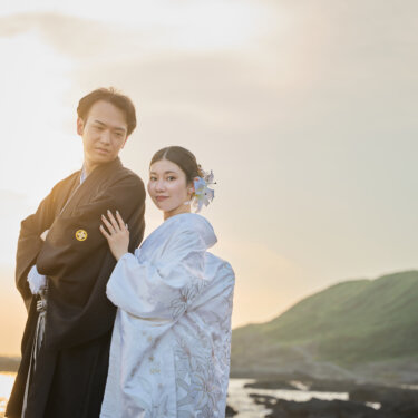 夕日が海を照らす城ヶ島の磯で、寄り添ってこちらを見つめる紋付袴の新郎と白無垢の花嫁。サンセットの和装前撮り