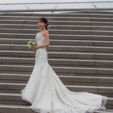 木のデッキの階段にレースのトレーンを広げて立つ、マーメイドドレスの花嫁のソロカット。小さなブーケを手にした洋装前撮り