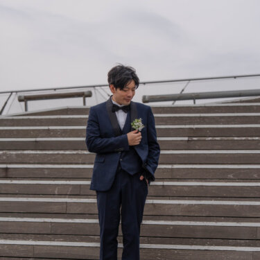 大さん橋の木の階段でブートニアに手を添えて立つ、ネイビーのタキシードの新郎のソロカット。洋装前撮りの新郎ショット