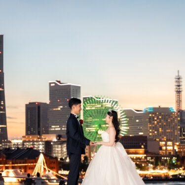 夕焼けに染まるみなとみらいの観覧車と赤レンガ倉庫を背景に、向かい合って見つめ合う新郎新婦の横浜の夜景前撮り