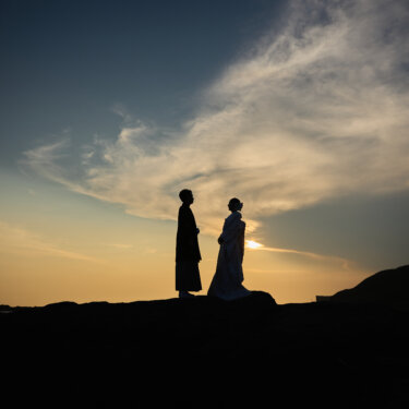 夕焼けの空の下、岩場の上で向かい合う紋付袴の新郎と白無垢の花嫁のシルエット。海辺のロケーションフォト