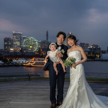 夜景が灯り始めた海を背に、赤ちゃんを抱いて並ぶ新郎新婦の家族写真。大さん橋で撮る子連れの前撮り