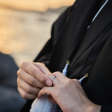 夕暮れの海を背に、紋付袴の新郎が結婚指輪をはめた手を重ねるアップ。和装前撮りで残す指輪のディテールカット