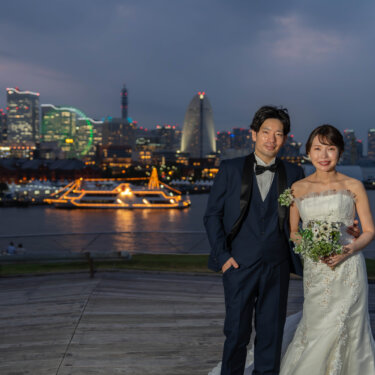 夕暮れのみなとみらいの夜景を背に大さん橋のデッキに並ぶ、ネイビーのタキシードの新郎とマーメイドドレスの花嫁。横浜の洋装ロケーションフォト