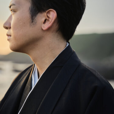 夕暮れの海を背に遠くを見つめる紋付袴の新郎の横顔。家紋が映える和装前撮りの新郎ソロカット