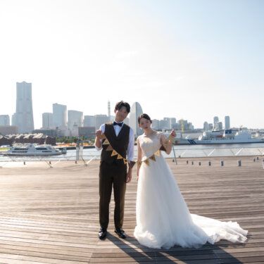 ガーランドを手に並んで立つ新郎とロングトレーンの花嫁。みなとみらいの街並みを望む大さん橋の屋上広場で撮る結婚写真