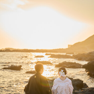 夕日にきらめく城ヶ島の海を前に、岩に並んで腰を下ろす紋付袴の新郎と白無垢の花嫁の後ろ姿。サンセットの和装前撮り