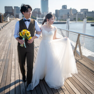 ひまわりのブーケを手に腕を組んで歩く新郎と、微笑むウェディングドレスの花嫁。海沿いの木のデッキで撮るナチュラルな前撮り
