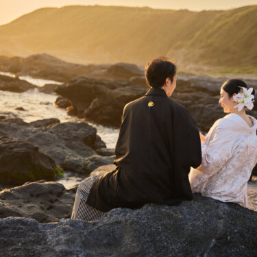 夕方の光に包まれた磯の岩に座り、語り合う紋付袴の新郎と白無垢の花嫁。城ヶ島の海辺で撮る和装フォトウェディング