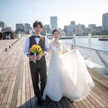 ひまわりのブーケを持つ新郎と、ドレスの裾をつまんで並んで歩く花嫁。大さん橋のウッドデッキで撮る夏の洋装前撮り