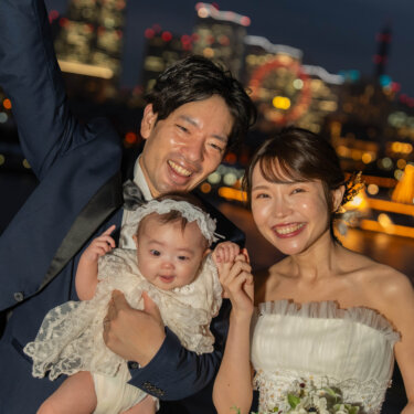 みなとみらいの観覧車の夜景を背に、ニコニコ笑う赤ちゃんを抱いて並ぶ新郎新婦。横浜の夜景で撮る家族写真の前撮り