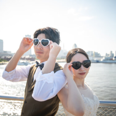 白と黒のハートのサングラスをかけて肩を並べる蝶ネクタイの新郎と花嫁。海辺の青空の下で撮るおしゃれなウェディングフォト