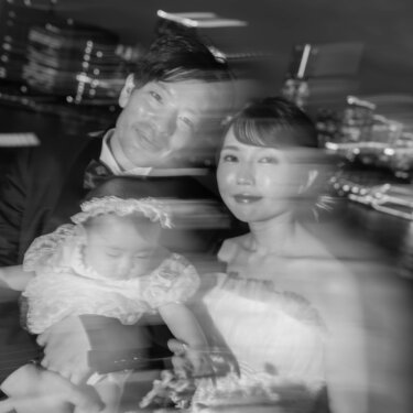 夜景の光が流れるように写ったモノクロの一枚で、赤ちゃんを抱いて笑う新郎新婦。夜に撮るアートな家族写真の前撮り