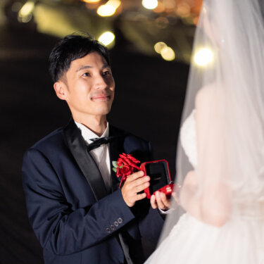 夜景のボケをバックに、赤いバラの花束とリングボックスを差し出してプロポーズする新郎のウェディングフォト