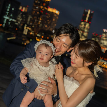 みなとみらいの夜景を背に、赤ちゃんを高く抱き上げて笑う新郎と寄り添う花嫁。家族で撮る夜景のフォトウェディング