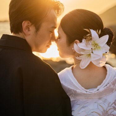 沈む夕日を背に、おでこを寄せ合う紋付袴の新郎とユリの髪飾りの白無垢の花嫁。逆光を生かした和装前撮りのポーズ