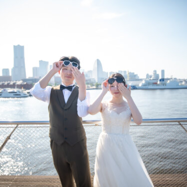 ハートのサングラスを掲げておどける新郎新婦。横浜港とランドマークタワーを背景にした、遊び心のある前撮りアイテムのポーズ