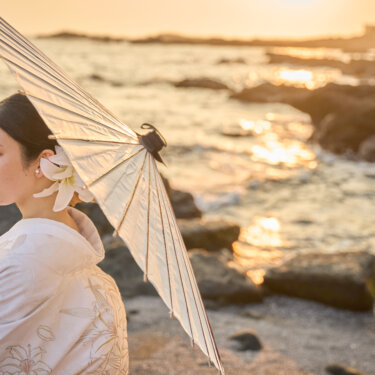 夕日がきらめく城ヶ島の磯で、和傘を肩にかけて振り返る白無垢の花嫁。海辺で撮る和装前撮りの花嫁ソロカット