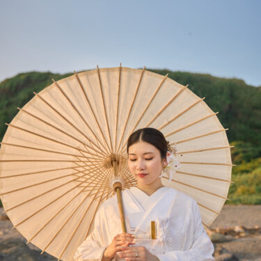 緑の丘と青空を背に、和傘を差して目を伏せる白無垢の花嫁。城ヶ島の海岸で撮る和装ロケーションフォト