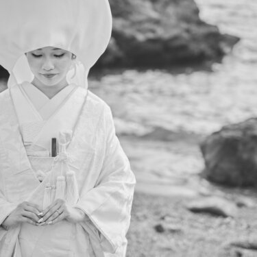 岩場の海辺で綿帽子をかぶり、手を重ねて目を伏せる白無垢の花嫁。モノクロで仕上げた和装の結婚写真
